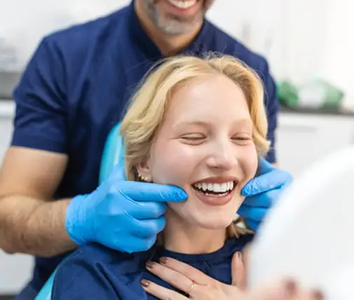 A person seated in a dental chair while another person wearing blue medical gloves gently positions hands near the person’s cheeks; a dental clinic environment is visible with bright lighting and medical attire.