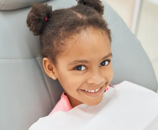 A person seated in a padded dental chair, wearing a light-colored protective bib, with hair styled into two small puffs, facing the camera with teeth visible; the background shows a clean clinical setting with neutral tones.
