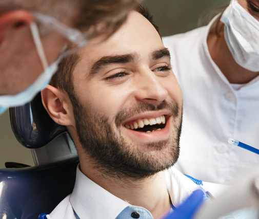 A person reclined in a dental chair with blue protective clothing visible, while two masked medical professionals stand nearby; one holds a dental instrument, and the clinical setting includes bright lights and dental equipment.