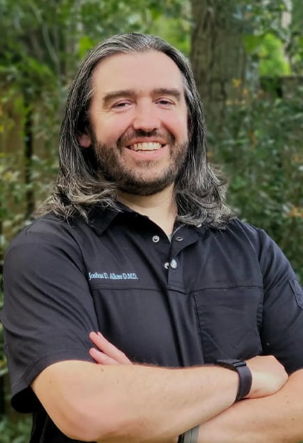 Joshua Allore, DMD, MS stands outdoors with arms crossed, wearing a black collared shirt, positioned in front of a leafy, green background with soft natural lighting.