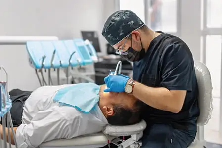 Dental clinician wearing protective gloves and mask performing a procedure on a person reclined in a dental chair, with equipment visible in the background.