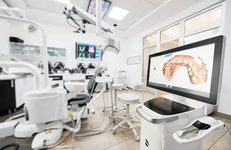 Modern dental operatory with treatment chairs and equipment; a monitor displays a digital 3D model of teeth on a cart-mounted screen.