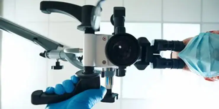 Dental operating microscope mounted on an adjustable arm, with gloved hands adjusting the controls in a clinical treatment room.