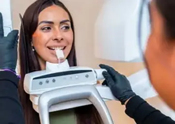 A person seated in a dental clinic using an intraoral scanning device positioned at the mouth, while a clinician wearing black gloves operates the scanner.